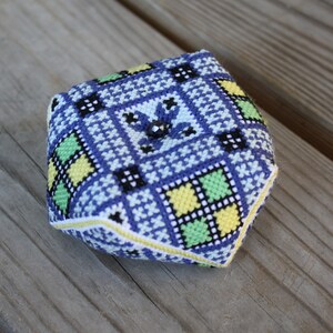 May include: A small, blue, yellow, and green patterned fabric cube with a flower design in the center.