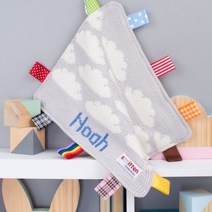 May include: A grey knitted baby blanket featuring white cloud patterns and colourful ribbon tags. The name "Noah" is embroidered in blue. The blanket has a tag that says "smitten".