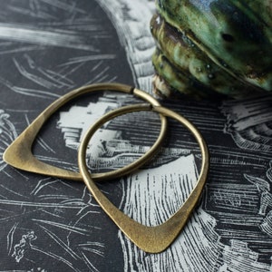 May include: Two brass hoop earrings with a unique, asymmetrical, angular design. The earrings are on a black and white patterned surface.