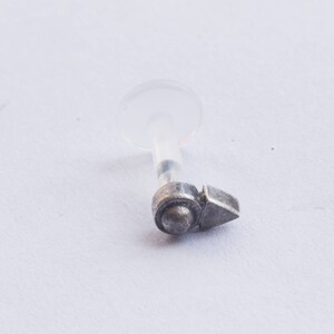 Puede incluir: Joyer&iacute;a corporal de plata con un respaldo de pl&aacute;stico transparente y un peque&ntilde;o amuleto en forma de flecha.