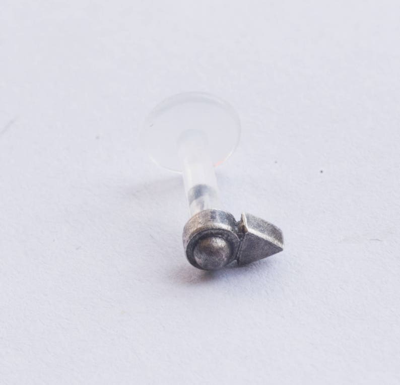 Puede incluir: Joyer&iacute;a corporal de plata con un respaldo de pl&aacute;stico transparente y un peque&ntilde;o amuleto en forma de flecha.