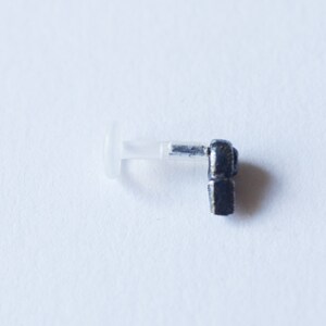 Puede incluir: Un piercing de cuerpo de metal negro con un respaldo de pl&aacute;stico transparente. El piercing tiene la forma de una peque&ntilde;a figura estilizada.