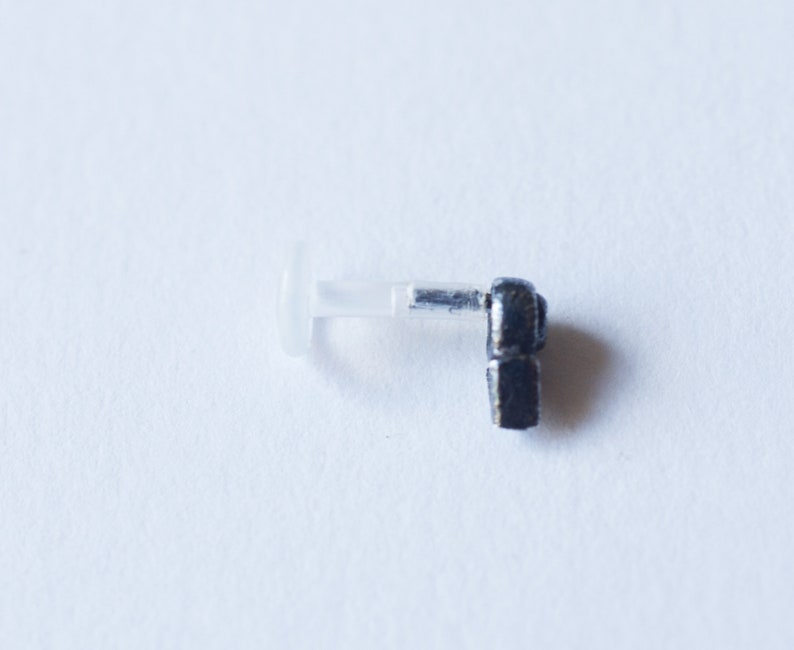 Puede incluir: Un piercing de cuerpo de metal negro con un respaldo de pl&aacute;stico transparente. El piercing tiene la forma de una peque&ntilde;a figura estilizada.