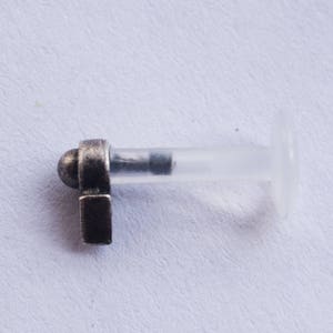 Puede incluir: Una pieza de joyer&iacute;a corporal de plata con una barra de pl&aacute;stico transparente y un tap&oacute;n decorativo. El tap&oacute;n tiene una parte superior redondeada y una base rectangular.