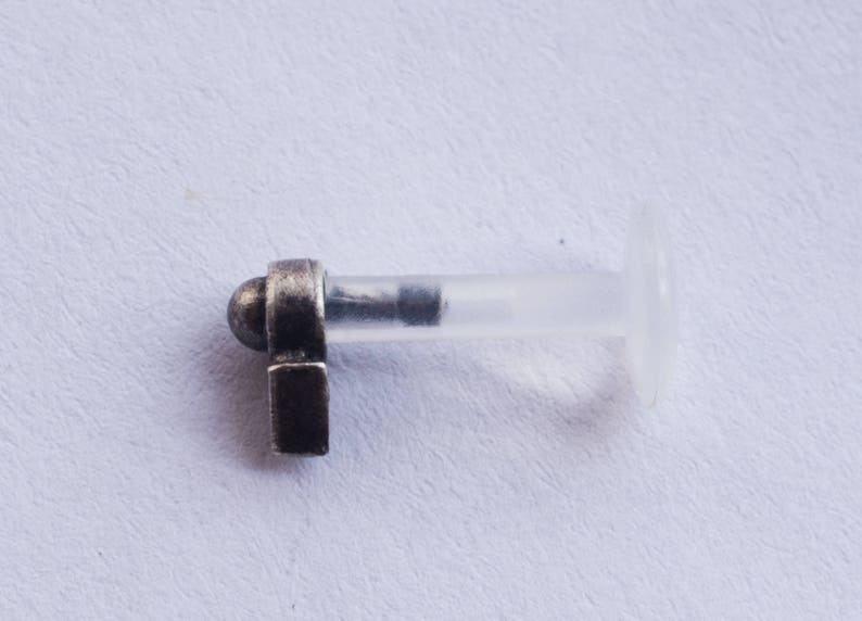 Puede incluir: Una pieza de joyer&iacute;a corporal de plata con una barra de pl&aacute;stico transparente y un tap&oacute;n decorativo. El tap&oacute;n tiene una parte superior redondeada y una base rectangular.