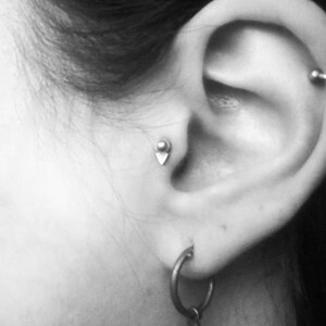 Puede incluir: Primer plano de la oreja de una persona con dos piercings. El piercing inferior tiene un peque&ntilde;o pendiente de plata, y el piercing superior tiene un aro de plata.