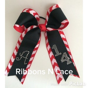 May include: A black and red chevron patterned hair bow with a white elastic band. The bow has rhinestones spelling out "14" on one side and "SA" on the other side. The bow is tied with a red ribbon and has the text "Ribbons N Lace" printed below it.