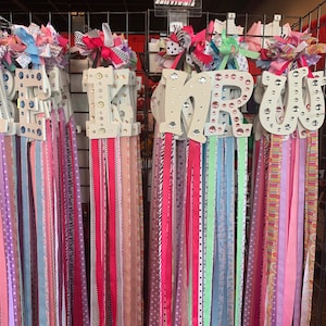 May include: A display of wooden letters with colorful ribbons attached. The letters are decorated with rhinestones and spell out the word "FREAKIN". The price tag reads "Large Anthropologie Style Hair Bows $15.00".
