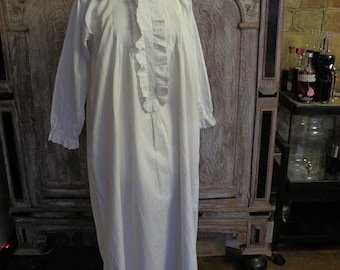 May include: A white cotton nightgown with a ruffled collar and long sleeves. The nightgown is displayed on a wooden stand against a carved wooden cabinet.