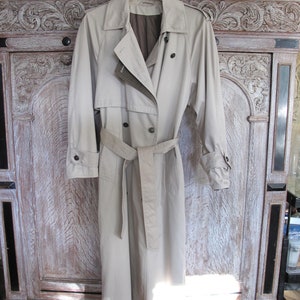 May include: A beige trench coat with a double breasted closure, a belt, and long sleeves. The coat is hanging on a hanger against a whitewashed wooden background.