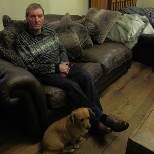 May include: A man wearing a gray and white sweater sits on a brown leather couch with a small brown dog. The couch has several brown and gray throw pillows. The man is wearing blue jeans and brown shoes. The dog is sitting on the floor in front of the couch.