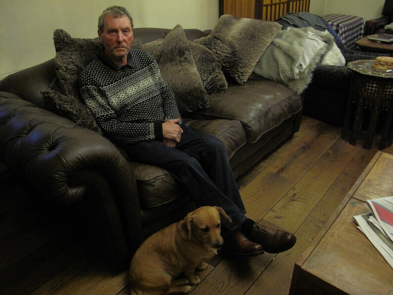 May include: A man wearing a gray and white sweater sits on a brown leather couch with a small brown dog. The couch has several brown and gray throw pillows. The man is wearing blue jeans and brown shoes. The dog is sitting on the floor in front of the couch.