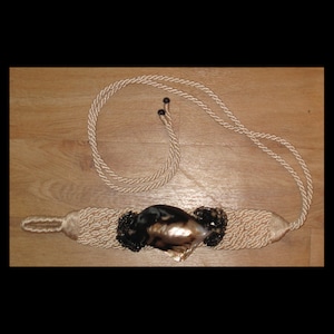 May include: A beige rope tassel with a large black and brown seashell accent. The tassel is tied in a knot and has a loop at the end.