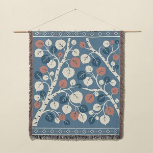 May include: A woven wall tapestry featuring a blue background with a pattern of white birch tree branches and leaves in shades of white, dark blue, and muted orange. The rectangular textile has a fringed edge and a geometric border.