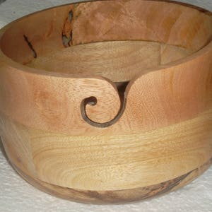 May include: A wooden yarn bowl with a natural wood finish and a carved yarn guide. The bowl is made of light brown wood with darker brown streaks.