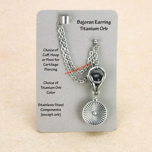 Könnte beinhalten: Silberfarbener Bajoran-Ohrring mit Titan-Kugel und Kette, die mit einer Manschette verbunden ist. Der Ohrring hat einen kreisförmigen Anhänger mit Sonnenstrahl-Design. Text auf der Karte: "Bajoran Earring Titanium Orb".