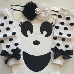 May include: A white baby bodysuit with a large black ghost face design, paired with matching white leg warmers with black polka dots and black bows. A black and white floral headband with a pearl center completes the set.