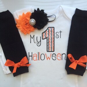 May include: A white baby bodysuit with the text "My 1st Halloween" in black and orange. The number "1" has an orange and black zig-zag design. Black leg warmers with orange bows and a floral headband complete the outfit.