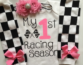May include: A white baby onesie with the text "My 1st Racing Season" and a large pink number 1. The outfit includes black and white checkered leg warmers with pink bows and a pink floral headband.