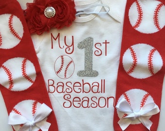 May include: A white baby onesie with the text "My 1st Baseball Season" in red and silver glitter. Red leg warmers with baseball graphics and white bows. A red flower headband with a pearl center.