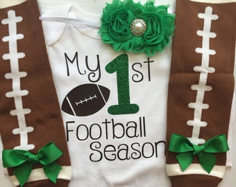 Puede incluir: Un body de bebé blanco con el texto "My 1st Football Season" y un gráfico de fútbol americano. Incluye calentadores de piernas marrones y blancos con temática de fútbol americano con lazos verdes y una diadema de flores verdes.