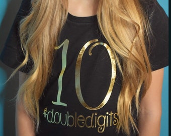 May include: A black t-shirt with the number 10 in gold foil and the hashtag #doubledigits. The shirt is worn by a person with long blonde hair, against a blue background.