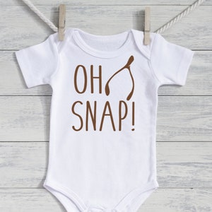 May include: White baby onesie with short sleeves, featuring the phrase "OH SNAP!" in brown lettering and a brown wishbone graphic. The onesie is hanging from a clothesline with a wooden clothespin.