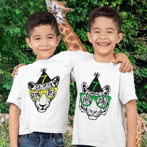 May include: Two boys wearing white t-shirts with animal face graphics and the number 7 on party hats. One shirt features a leopard with yellow glasses, the other a tiger with green glasses. Both are smiling in front of a green background.