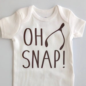 May include: A white baby onesie with short sleeves and a round neckline. The words "OH SNAP!" are printed in dark brown, along with a wishbone graphic. The onesie has snap closures at the bottom.