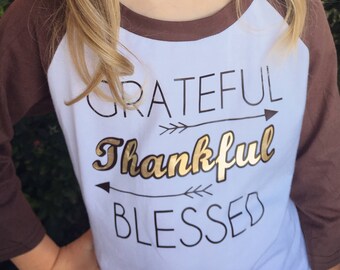 May include: A tan felt hat with heart-shaped cutouts and a gold glitter band. The person is wearing a white and brown baseball tee with the words "Grateful, Thankful, Blessed" in gold and black lettering.