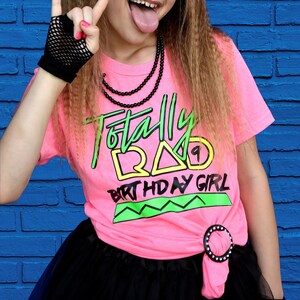 May include: A girl in a pink t-shirt with the words "Totally Rad Birthday Girl" in neon green and yellow. She wears a black net glove, a black beaded necklace, and a black bow. The background is a blue brick wall.