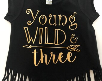 May include: Black t-shirt with gold text that reads "Young Wild & Three" with an arrow design. The shirt has a fringe detail at the bottom and a round neckline. The shirt is for a girl.