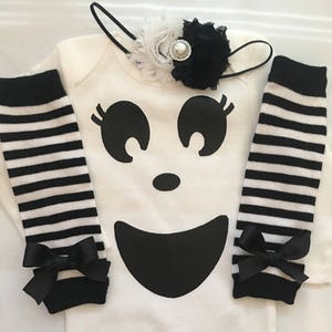 May include: A white baby bodysuit with a large black ghost face design, paired with black and white striped leg warmers and a matching headband. The leg warmers have black bows at the ankles.