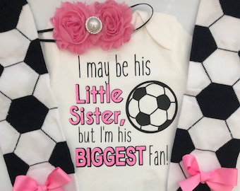 May include: A white baby bodysuit with the text "I may be his Little Sister, but I'm his BIGGEST Fan!" and a soccer ball graphic. Includes black and white soccer ball leg warmers with pink bows and a pink floral headband.