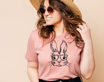 May include: A light pink t-shirt featuring a black and white illustration of a bunny wearing glasses. The bunny's glasses have a leopard print design. The person is wearing a straw hat and sunglasses.