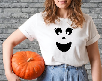 May include: White t-shirt featuring a black ghost face design with large eyes and a smiling mouth. An orange pumpkin is held in the image. The shirt is paired with blue denim jeans, creating a casual Halloween look.