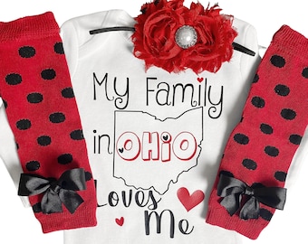 May include: A white baby onesie with the text "My Family in OHIO Loves Me" and a red heart graphic. The outfit includes red leg warmers with black polka dots and black bows, and a red flower headband.