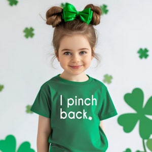May include: A young girl wearing a green t-shirt with the text "I pinch back" in white. She has a green bow in her hair and is wearing green pants. The background features shamrock decorations.