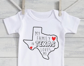 May include: White baby onesie with the text "MY FAMILY IN TEXAS LOVES ME" in black, with a Texas outline and two red hearts. The onesie is hanging from a clothesline with a wooden clip.