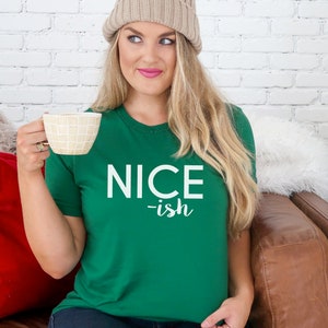 May include: A green t-shirt with the words "NICE -ish" printed in white. The t-shirt is being worn. The person is holding a beige mug. A red cushion and a Christmas tree are in the background.