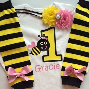 May include: A white baby bodysuit featuring a yellow number one and a cartoon bee design. The bodysuit is personalised with the name "Gracie" in pink. The set includes yellow and black striped leg warmers with pink bows and a floral headband.
