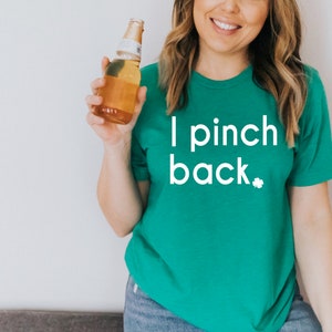May include: A green t-shirt with the text "I pinch back" in white, along with a small shamrock. The person is holding a beer bottle. The shirt is a solid color and has a round neckline.