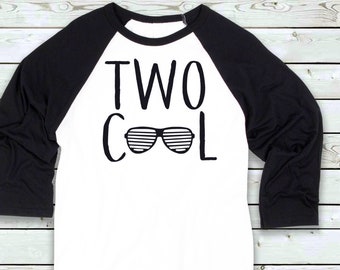 May include: White and black raglan style shirt with the words "TWO COOL" in black block letters and a pair of sunglasses. The sleeves are black, and the body of the shirt is white. The shirt is made of a soft material.