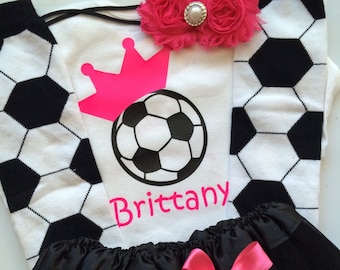 May include: A soccer-themed outfit featuring a white t-shirt with a soccer ball and crown graphic, the name "Brittany", and black and white soccer ball socks. A black tutu skirt with a pink bow and a pink floral headband complete the ensemble.