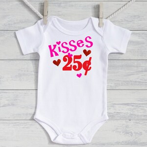 May include: White baby bodysuit featuring the text "Kisses 25p" in pink and red, with heart decorations. The garment is suspended from a clothesline against a white wooden backdrop.