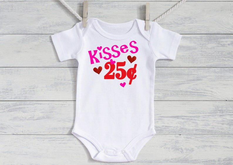 May include: White baby bodysuit featuring the text "Kisses 25p" in pink and red, with heart decorations. The garment is suspended from a clothesline against a white wooden backdrop.