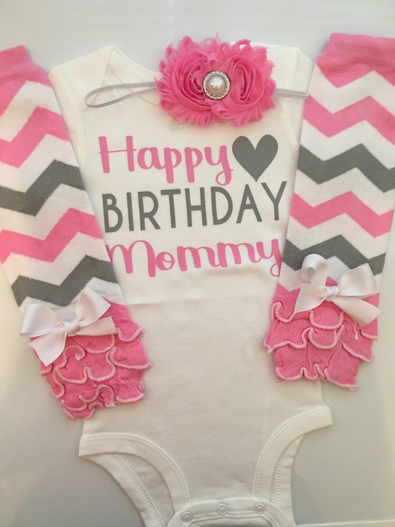 May include: A white baby bodysuit with the text "Happy BIRTHDAY Mommy" in pink and gray. The set includes pink and gray chevron leg warmers, a pink flower headband, and pink ruffled leg warmers with white bows.