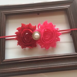 May include: A red and hot pink floral headband with a pearl-centred embellishment. The headband features two fabric flowers and a thin red elastic band. The flowers are set against a white background and framed by a brown wooden frame.