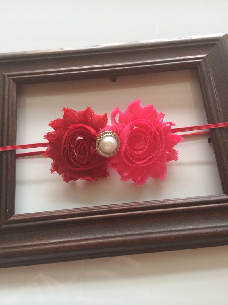 May include: A red and hot pink floral headband with a pearl-centred embellishment. The headband features two fabric flowers and a thin red elastic band. The flowers are set against a white background and framed by a brown wooden frame.