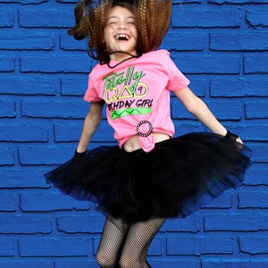 May include: A girl wearing a pink t-shirt with the text "Totally Rad Birthday Girl", a black tutu, fishnet stockings, and green leg warmers. Her hair is styled in a retro fashion, and she is jumping in front of a blue brick wall.